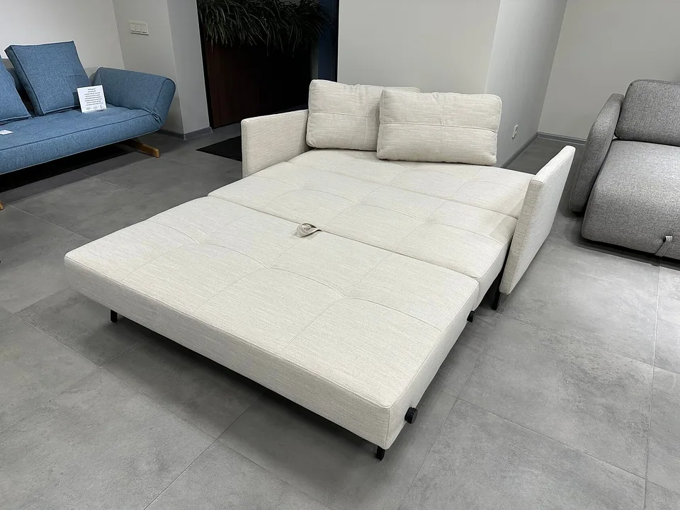 Cubed 140 with Arms разложен в кровать, в три четверти слева сверху: стёганое спальное место выдвинуто вперёд на чёрных ножках, на нём тканевая петля, спинки-подушки остались между подлокотниками