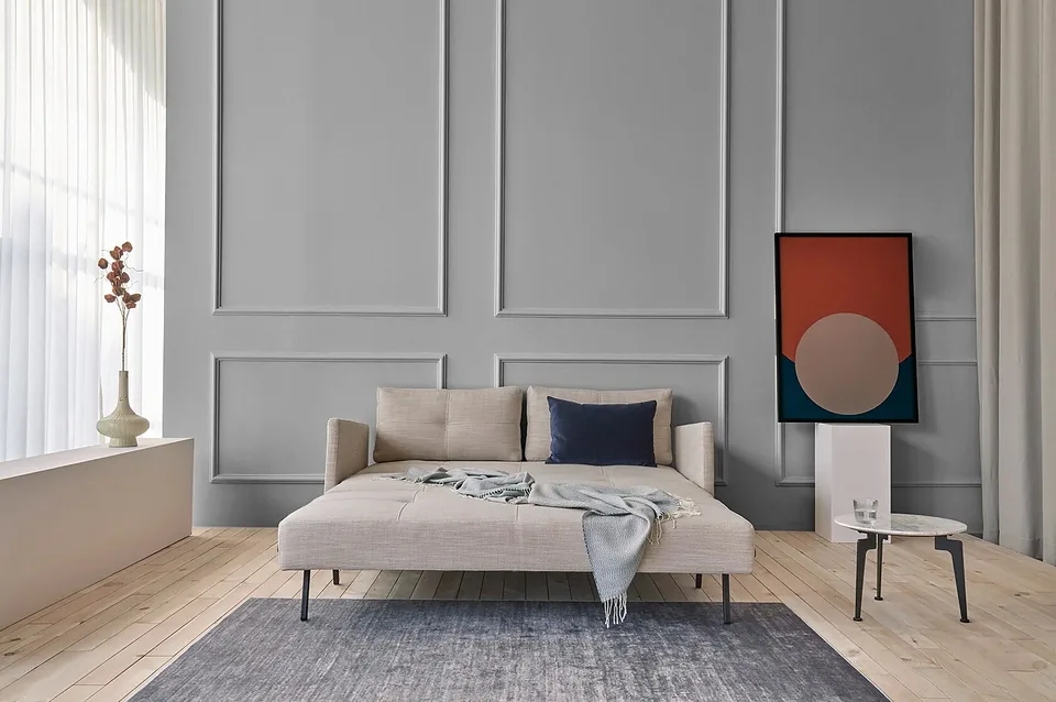 Cubed Sofa Bed With Arms в бежевой меланжевой обивке разложен в кровать, вид спереди: на спальном месте серо-голубой плед с бахромой, у спинки тёмно-синяя подушка