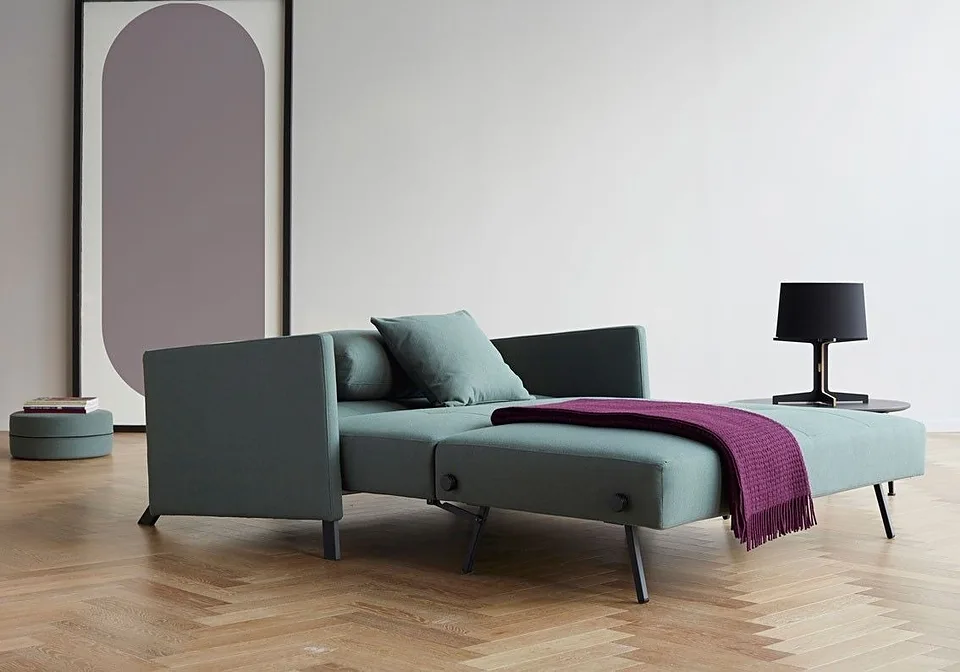 Cubed Sofa Bed With Arms в серо-зелёной обивке разложен в кровать, вид сбоку в три четверти: подлокотники на чёрных ножках, между ними подушка, на спальном месте фиолетовый плед с бахромой