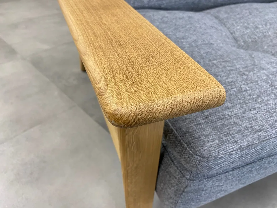 Левый дубовый подлокотник Dublexo Frej Oak крупным планом: скруглённый торец над передней ножкой и рисунок волокон, рядом стёганое сиденье в серой ткани