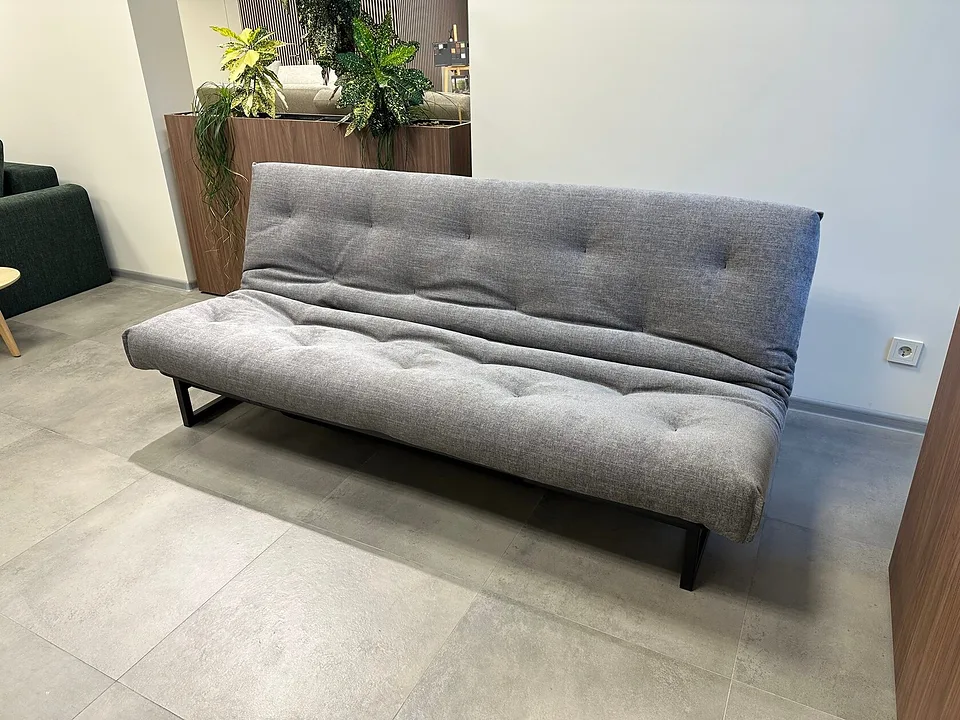 Диван-кровать Fraction 120, Innovation Living, в серой меланжевой ткани в салоне, вид спереди-слева: стёганые спинка и сиденье на низком чёрном металлическом каркасе, позади кашпо с растениями