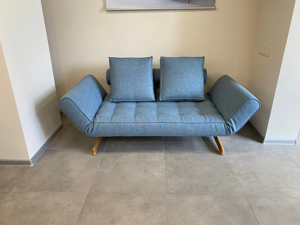 Кушетка Ghia Oak, Innovation Living, в светло-голубой обивке Mixed Dance Light Blue 525 в нише, вид спереди: оба подлокотника подняты в верхнее положение, у валика две подушки, стёганое сиденье на дубовых ножках
