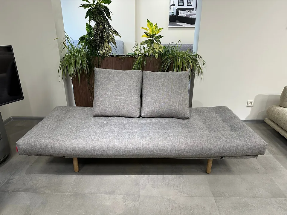 Кушетка-кровать Rollo, Innovation Living, в серой ткани 521 MIXED DANCE GREY в салоне, вид спереди: ровный стёганый матрас с опущенными боковыми частями, две подушки, светлые деревянные ножки, сзади кашпо с растениями